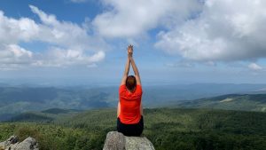 A woman in a red shirt relaxes on a mountain peak with arms raised, enjoying the scenic view.